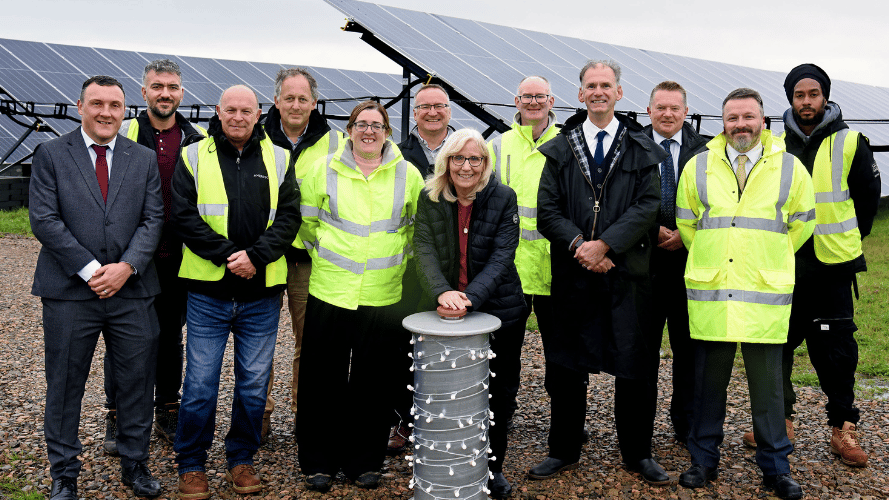 A dozen people standing outdoors, wearing a mixture of suits and high vis jackets. They are smiling into the camera, with one person - a woman - about to press a large button.
Rows of solar panels are in the background behind them.