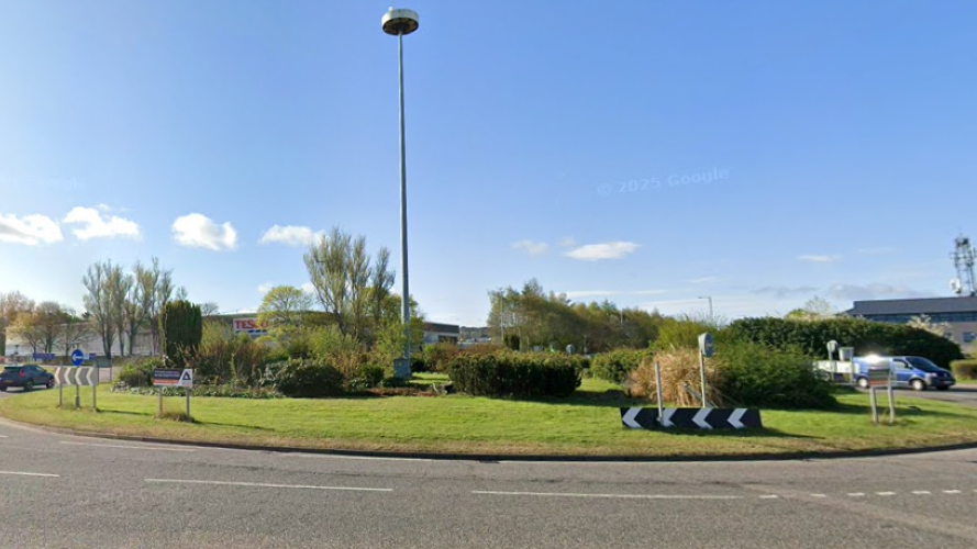 The roundabout is a large grassy feature with shrubs and road signs with black and white chevrons. There is a tall street light standing in the middle of the roundabout.