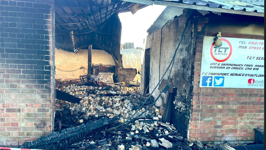 A burned out brick built barn, with a warped metal roof and charred masonry and bricks heaped on the ground