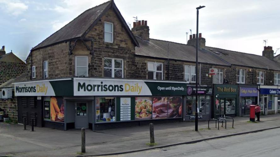 A row of shops at a junction between a main road and minor road, with a convenience store on the corner. It has a green and white sign with "Morrisons Daily" written in large green and yellow letters.