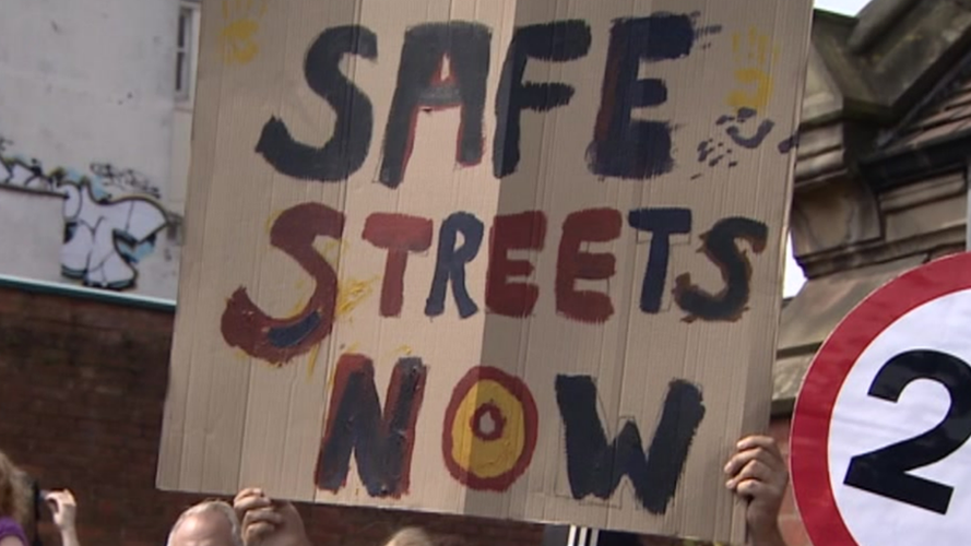 A brown cardboard sign with "safe streets now" written on it in black, red and yellow letters. It is being held up by someone during a protest. Part of a 20mph speed limit sign is on the right.