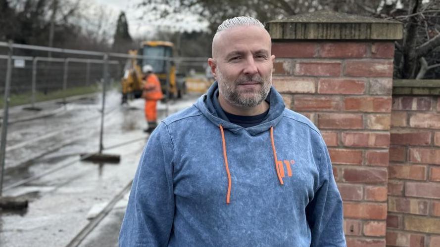 Image of Chris. He has a blue jumper on with an orange tinge on the front and on the toggles. He is stood in front of a Severn Trent worker in bright orange work gear behind some fencing. 