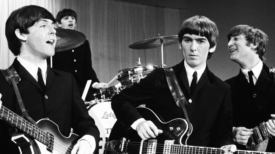 The Beatles in black and white, with Paul McCartney, Ringo Starr, George Harrison, and John Lennon dressed in suits playing their instruments.