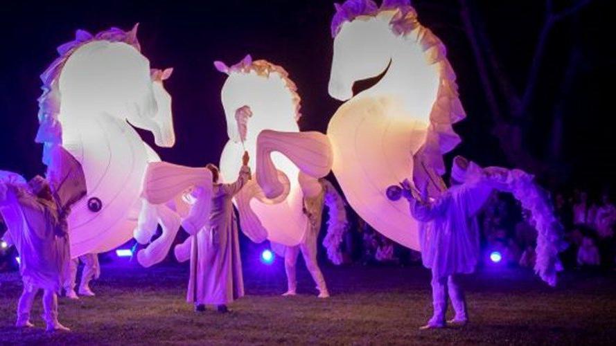 French street theatre artists wearing illuminated white and purple horse costumes in a field. 