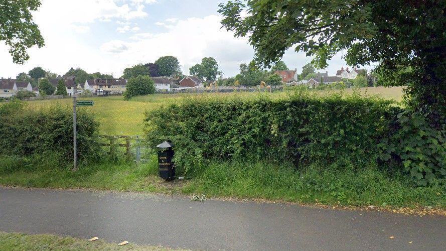 A public footpath sign pointing across a grassy field next to a metal gate and a black litter bin next to it. Houses are in the distance. 