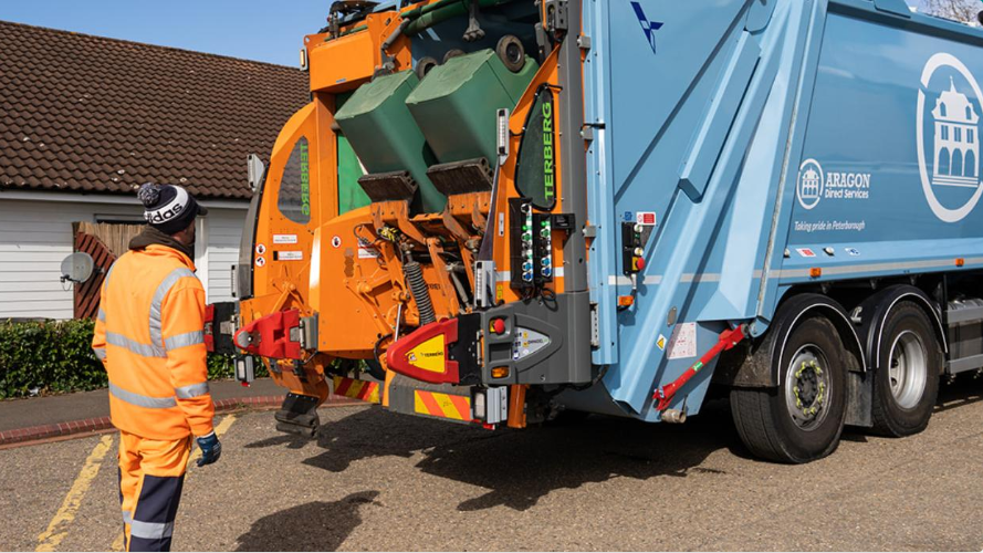 A bin collector wearing bright orange is standing behind a blue bin lorry, where green bins are being emptied into the back, outside someone's bungalow