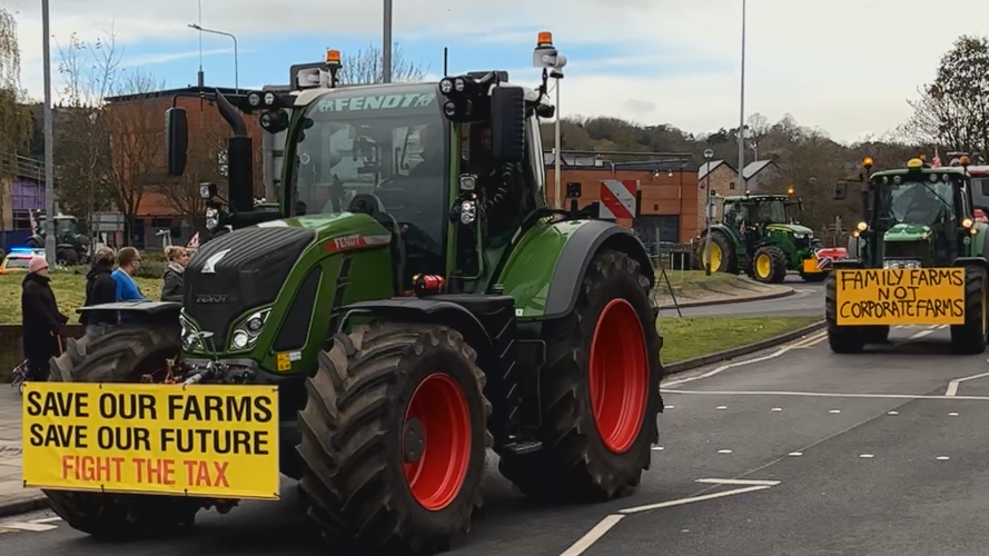 A green tractor travels toward the camera on the opposite side of a road. It has a yellow sign on its front that reads: "Save our farms. Save our future. Fight the tax." Another similar green tractor travelling behind it has a sign that reads: "Family farms. Not corporate farms."
