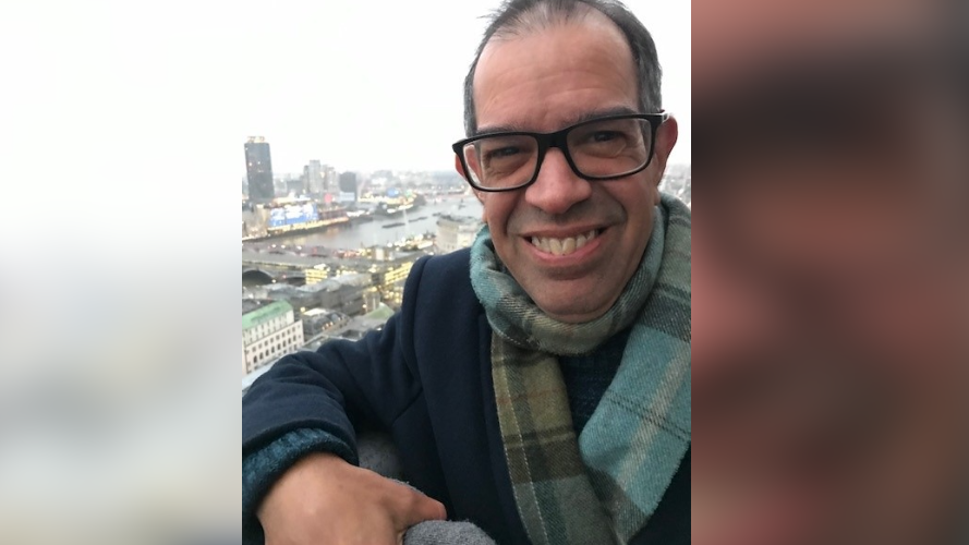 Andrew Wareham smiling at the camera in glasses and tartan scarf on a winters day from a balcony looking over a city