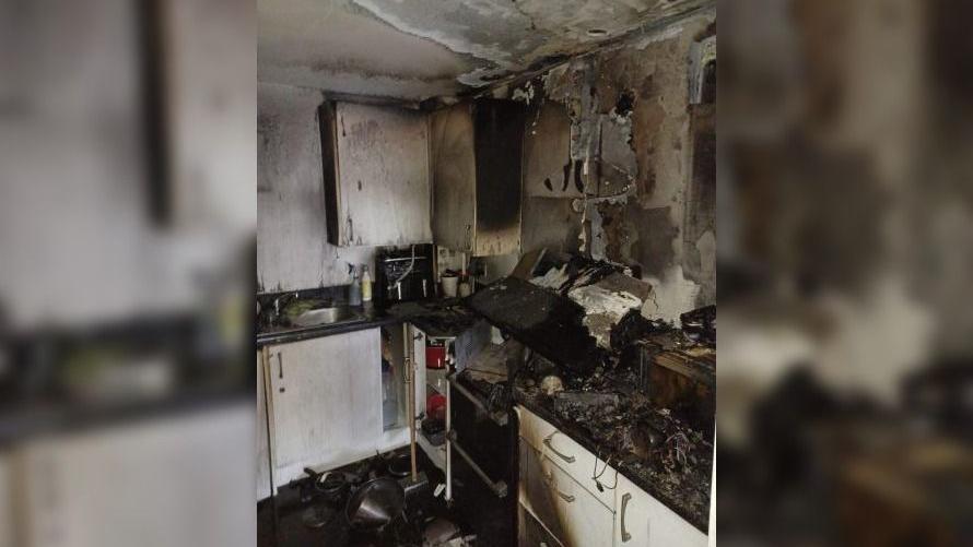 A kitchen which has melted worktops and is covered in black soot. The cooker hood has collapsed and fallen off the wall and on to the hob below. The white walls are grey with soot.