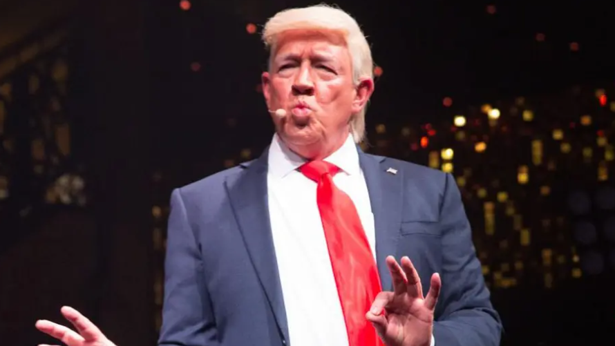 Mike Osman dressed as Donald Trump. He is wearing a blonde wig and pursing his lips together. He is wearing a blue suit and red tie.