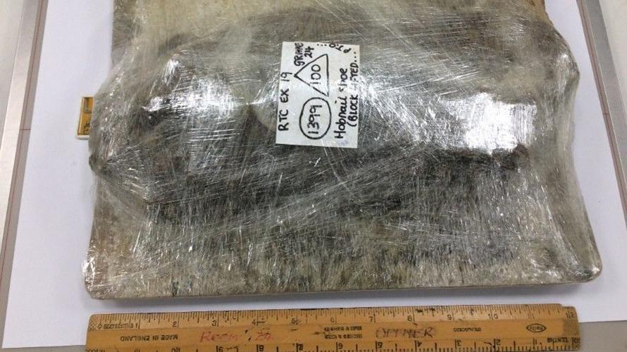 A clump of dirt wrapped in multiple layers of cling film on a blank surface. There is a white label on it and it lies next to a wooden 30cm ruler.