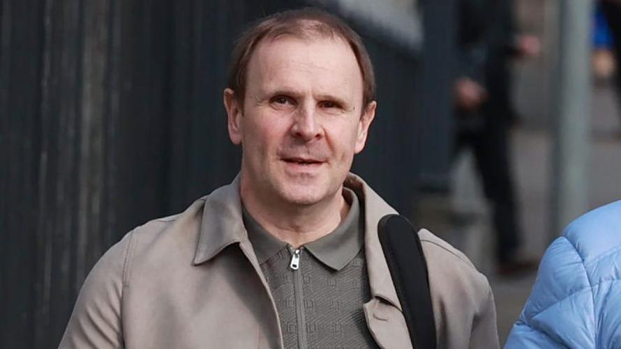 Winston Irvine, a brown-haired man, photographed outside court. He is pictured from the chest up amd wears a olive green zipped up thin jumper under a beige coat. A bag strap is visible on his shoulder.