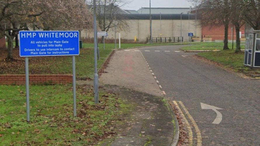 The exterior of the red-brick Whitemoor Prison. A sign on the left has a blue background with white text reading "HMP Whitemoor".