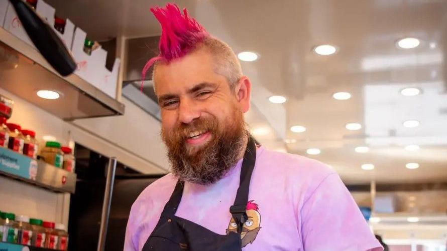 A man with a beard and pink mohawk hair is wearing a pink T-shirt and black apron. He is smiling and standing in a brightly-lit room with herb and spice jars on shelves next to him.