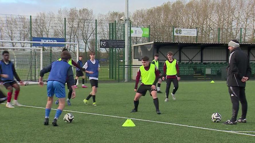 A group of young teenage boys, some in blue bibs and some in green ones, play football as a male adult coach stands to one side watching.