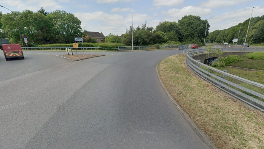 A junction at a road with a barrier either side. In the distance you can see some houses and few cars on the road. 