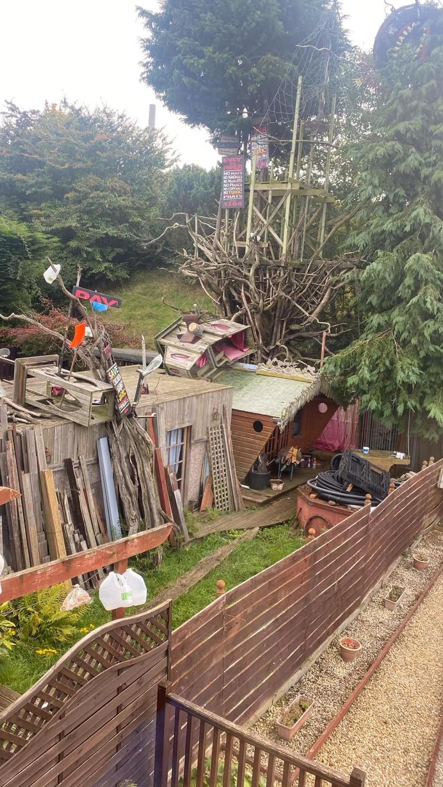 The remains of a galleon shaped boat in a tree in the background. A lot of wood and other items lie on the grass and along the fence. 