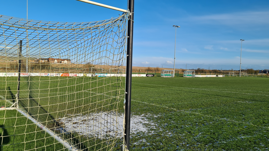 An empty football pitch with two very small areas for players to sit and some lights. There is a patch of frost in front of the goal post.