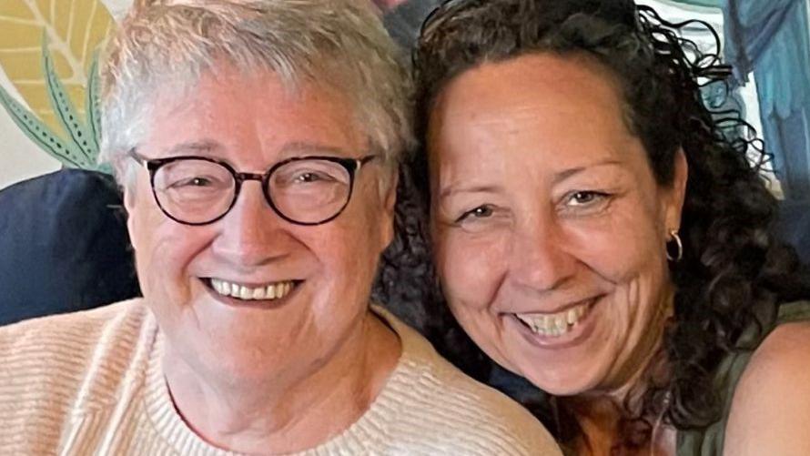 Susan sitting on a sofa with her daughter Emma, a middle-aged woman with long dark hair. They are both grinning at the camera