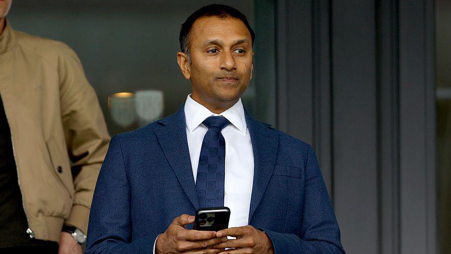 Shilen Patel watches a West Bromwich Albion game, wearing a suit and tie and holding a mobile phone
