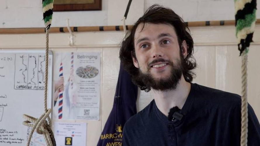 Iwan Cotgreave stood in a bell tower between two ropes with a bell ringing poster behind him.