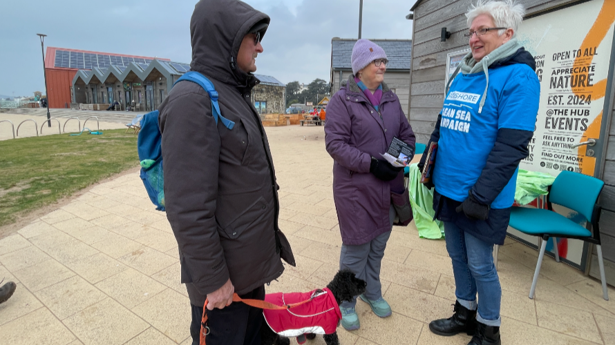 Three people stand on a pavement, one with a small black dog. They are dressed in cold weather gear and one is wearing a blue T-shirt with sme words on it.