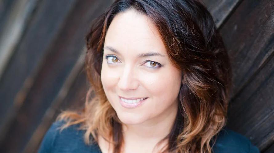 A close-up head and shoulders picture of Melissa Thom. She has hazel eyes and dark brown hair at her roots, which fades out in a balayage effect to light brown at the ends. She is wearing a teal top and standing against a wooden fence, smiling at the camera