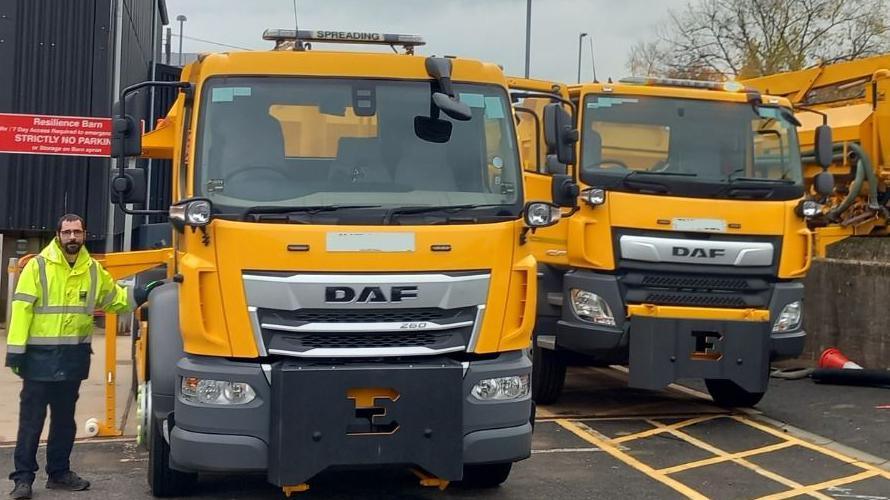 Two yellow DAF lorries parked up, facing the camera, with a council worker in hi-vis jacket standing beside