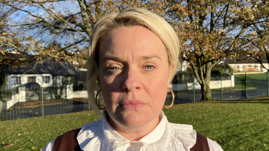 Principal of Drumahoe Primary School Emma Walker has blonde hair and is wearing large hooped earrings. She is wearing a white shirt with brown coloured jumper. She is standing in a grassy area near a large green gate and white building.