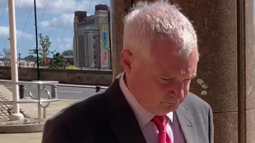 Timothy Gardner is wearing a suit and red tie. He has grey hair and is looking down at the ground while walking outside court. 