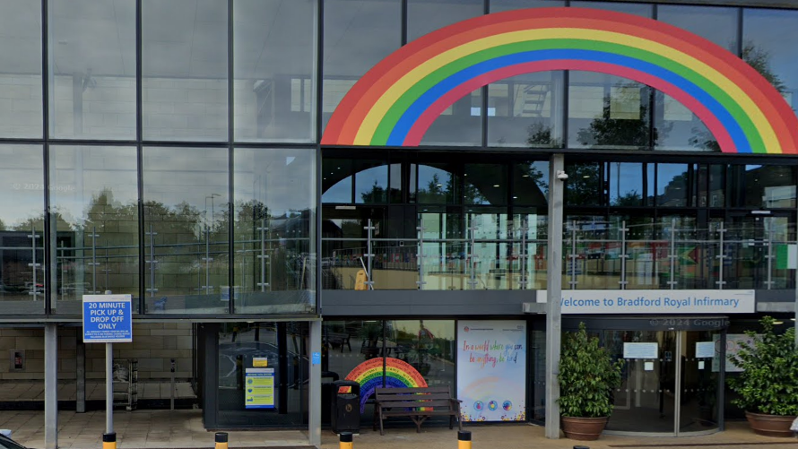 A glass fronted building with a revolving door on the right side. Above the door is a sign that reads Welcome to Bradford Royal Infirmary. A large rainbow decal stretches across part of the second floor. Outside the building there is a bench, two plants in pots and a sign that says 20 minute pick up and drop off only.