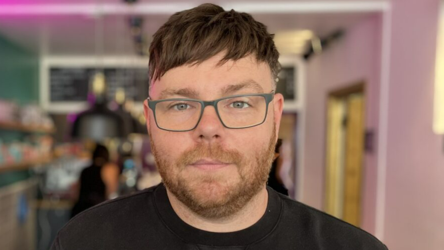 Dean Coles has short brown hair and wears glasses and a black sweater. He has a small smile on his face. Behind him is a menu on black chalkboard and heat lamps above a serving area.