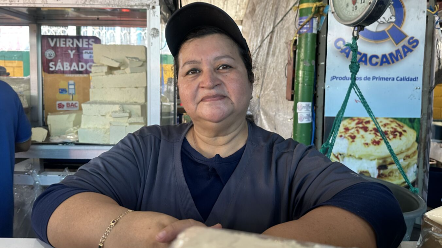 Nolvy Oriales is leaning on the counter at her cheese shop. She is wearing a dark baseball cap pushed back on her head. Behind her a scale is hanging from the ceiling and signs advertise products and list the opening days.