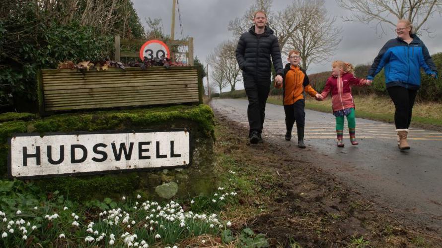 A family of four walk towards a Hudswell village sign holding hands