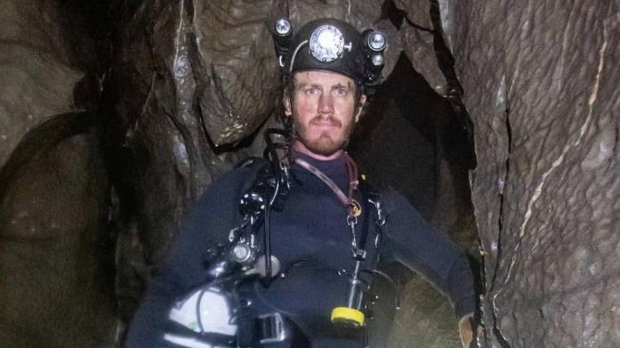 George Linnane inside a cave and looking at the camera. He is wearing a wetsuit and a safety helmet. 