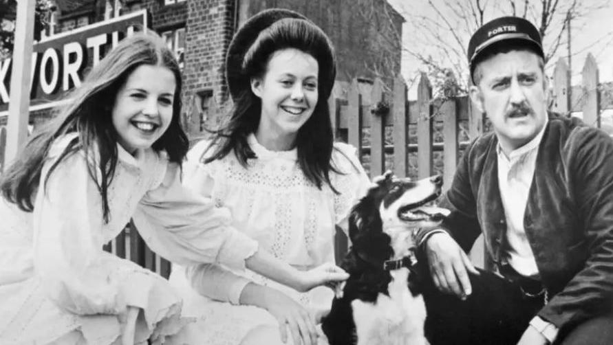 A black and white picture of the film version of the Railway Children showing two children on the left and a man on the right with a dog in the middle.