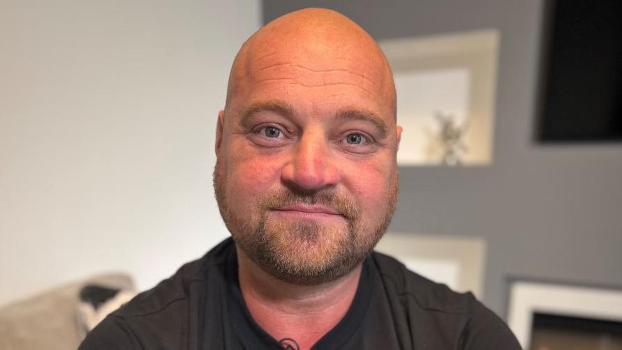 Offshore worker Phil Perry, who has a bald head and is wearing a black T-shirt, is smiling at the  camera. He has tattoos on arms.