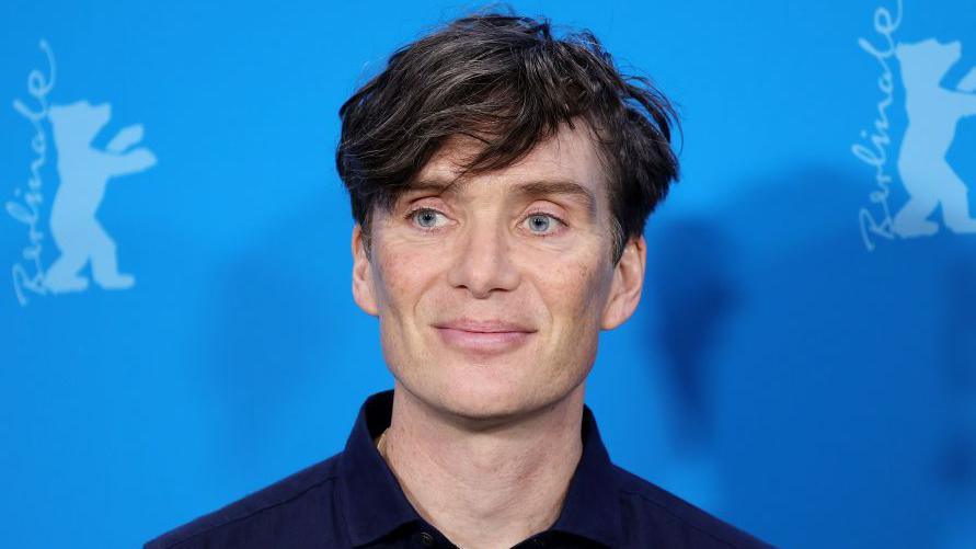 Cillian Murphy wearing a navy shirt smiling against a blue backdrop 