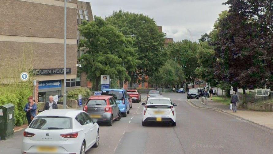 Rouen Road, Norwich, with several cars on the road, people walking up and down the street, trees are along the road, and a large brick building to the right.