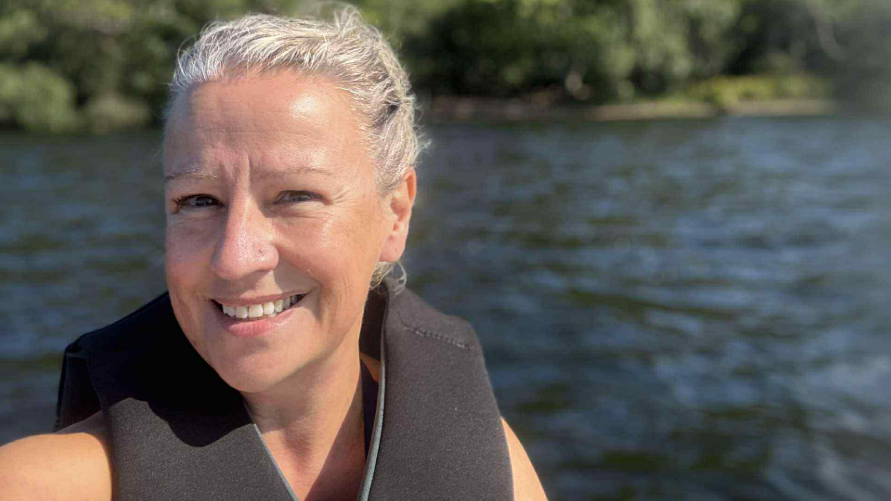 A selfie of Ellie smiling into the camera. She has her hair tied back and is wearing a black life jacket. Behind her is a dark body of water and trees.