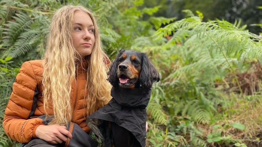 Lily-May pictured looking away from the camera, sat on a bench with her dog Sadie. She's wearing an orange waterproof coat and Sadie is wearing a black dog raincoat. Behind Lily-May is a bank of ferns and trees.