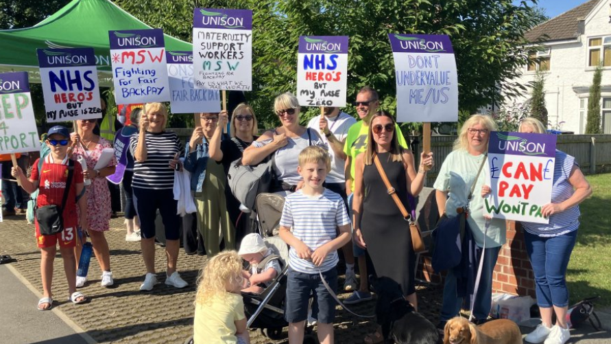 Maternity staff wearing casual clothes, some with children, hold placards with handwritten slogans including "can pay, won't pay" and "don't undervalue me/us".