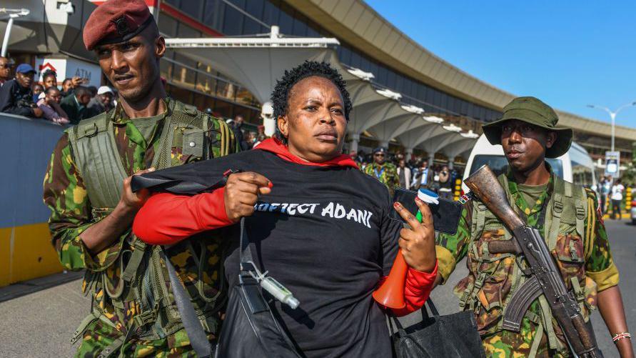 At the airport, two armed soldiers in military uniforms escort a female protester holding a vuvuzela and dressed in a black T-shirt that says "Adani"