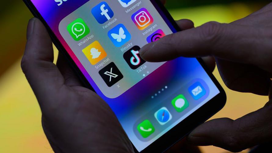 A close-up image of a phone in someone's hand. It has social media apps on the screen and a finger is going to tap on TikTok.