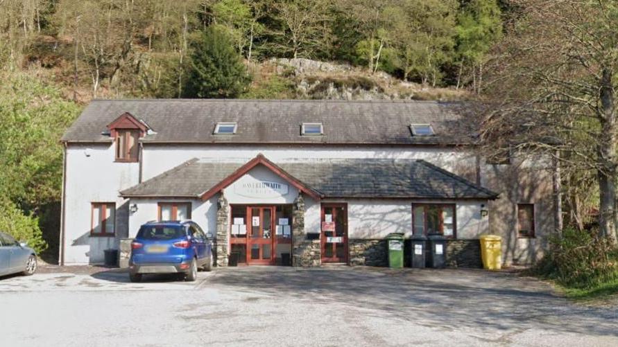 The Haverthwaite Surgery. It is a white building with wooden doors and windows. It has trees behind it and a car park in front.