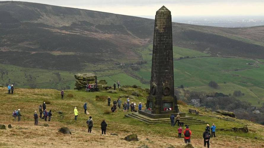Scores of people gather while socially distancing during a memorial event in 2020 around the Saddleworth memorial. It is a tall slender obelisk pillar on a hill, surrounded by other hills in the Pennine countryside.