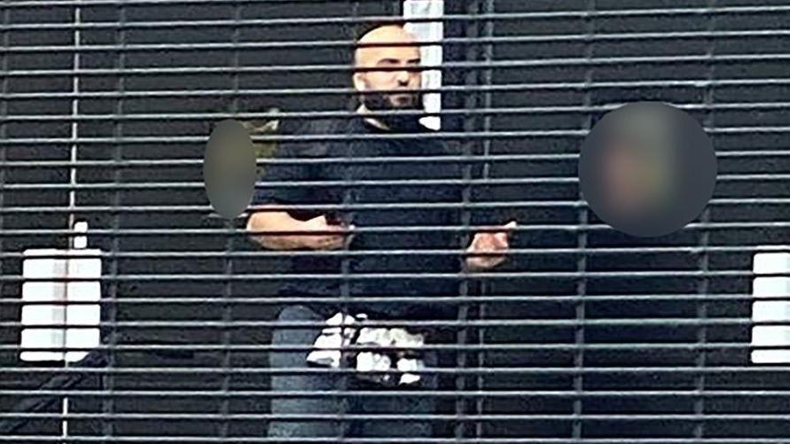 An image of a bald man with a beard in a black jumper and jeans seen through fencing holding his hands aloft. 