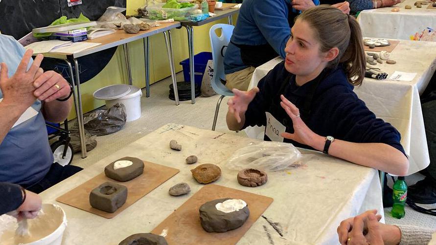 A woman speaking to a group of people around a table. On the table are some small clay casts filled with plaster