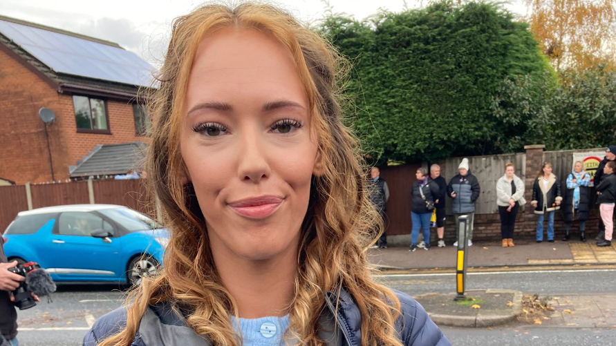 A girl with chestnut coloured hair and brown eyes stands in front of the roadside waiting for Keith's procession - she wears a blue cardigan and a navy blue jacket. Behind her is the crowd waiting for Keith's funeral procession