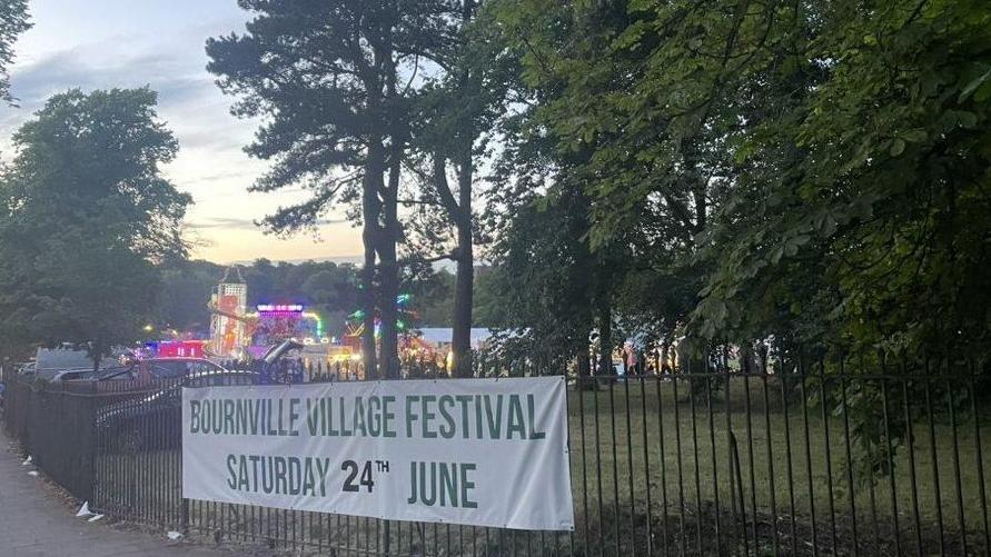 Fairground rides which include a helter skelter which is lit up, trees and a sign saying "Bournville Village Festival".